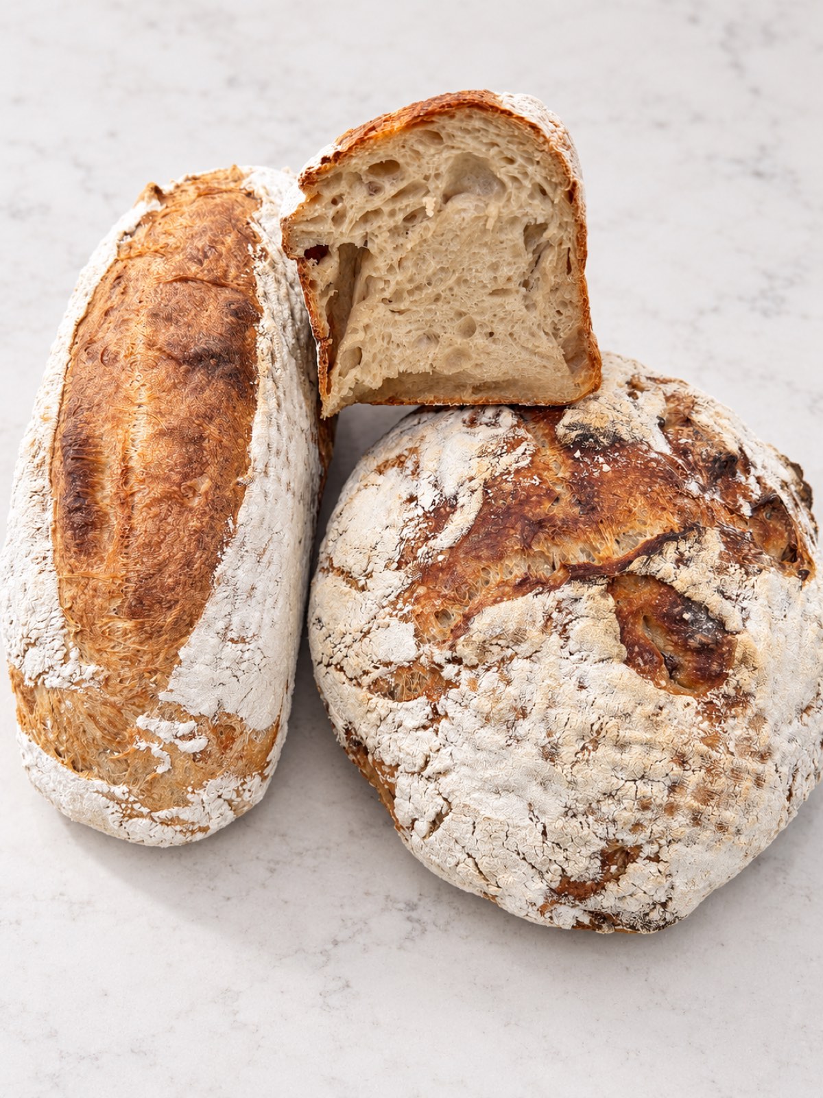 A long Double-Fed Sweet Levain, a round Country Blonde with Pecan, and a cut piece showing the open crumb, on a marble counter.